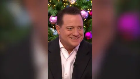 Brendan Fraser, in a white shirt and dark jacket, smiles in front of a Christmas tree