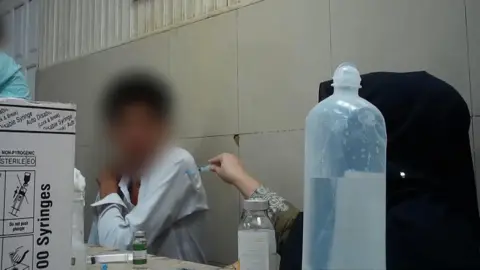 A scene from a hospital. A woman, whose face is obscured, is injecting a patient, his face blurred. She is injecting him without wearing gloves and putting the needle through the sleeve of his shirt.