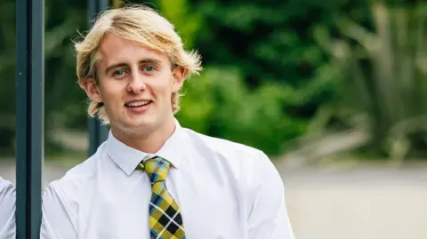 A young man with blonde hair is smiling casually. He's leaning against a window and is wearing a white shirt with a yellow and black checked tie. 