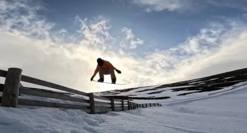 William Warnock A person on a snowboard performing a jump over a wooden snow fence on a snowy slope. The sky above is bright with scattered clouds, and the hillside is partially covered in snow.