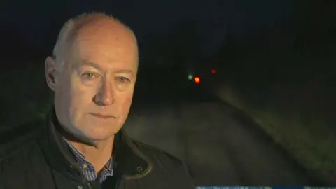 A man standing on a dark road. He is bald and is wearing a blue shirt and a dark coloured jacket.