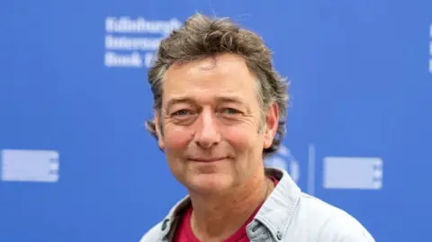 Getty Images Ned Boulting smiles at the camera during a book festival. He has short brown and grey hair. He is wearing a blue shirt with a red t-shirt underneath. He is standing in front of a blue background.