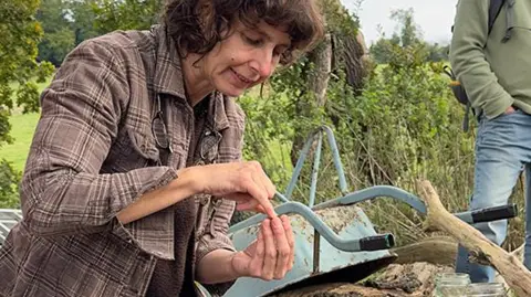 Deb Roberts Tara Downs sat in nature, looking at something in her hand. Behind her a man is standing looking on. We cannot see his face. Tara is wearing a brown flannel shirt and sat next to a light blue wheel barrow.