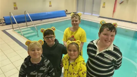 BBC Five pupils from La Rondin school were standing in front of a swimming pool. four boys and a girl in the center. They all wear a pair of yellow Pudsey ears