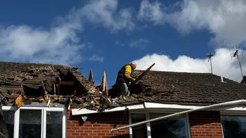 A man in a yellow top stands in a large hole in the roof of two houses