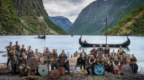 Norway players in Viking dress and with weapons and shields pose in front of a fjord with Viking boats