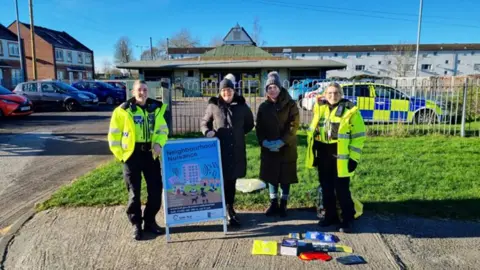 Hull City Council Two female police officers in high-vis uniforms stand either side of two council workers in dark coats and hats. In front of them is a sign saying "Neighbourhood Nuisance". In the background are houses and a police car.