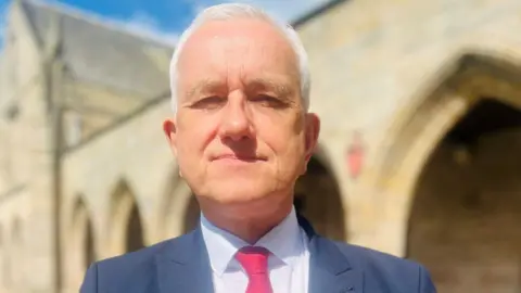 BBC Prof Peter Edwards, a man with white hair and wearing a blue suit jacket, light shirt and red/pink tie, in a university setting.