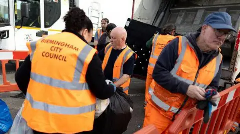 Residents hand rubbish to refuse workers at a Mobile Household Waste Centre in Senneleys Park on 8 April, 2025. Men and women workers are wearing orange hi-vis jackets.