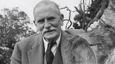 Getty Images A black and white photo of a man with short white hair and a white mustache. He is wearing a three piece suit with a white shirt and dark tie with lighter spots on it. He is resting one hand on a tree stump as he looks into the camera.