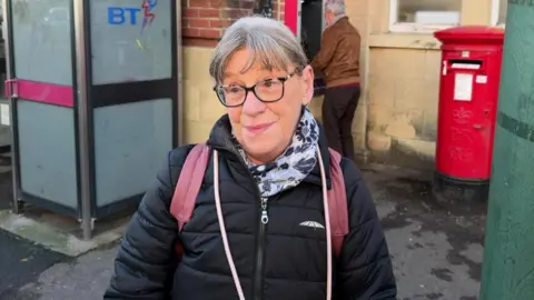 Jane Kempton stands outside the post office, there is an old BT phone booth to the left behind her and a red box box in the background to her right, a man is using a cash point near the post box too. Ms Kempton has grey and white hair, which is tied back and a fringe framing her face. She wears thick framed black glasses and is looking slightly off camera, wearing a blue floral scarf, a black puffer jacket and has dusky pink rucksack straps.