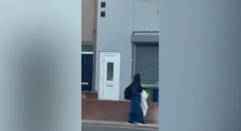 Facebook Still from a video showing a woman walking down a street carrying a white sheet. She is passing a property with a shutter over a window and a door to the side.