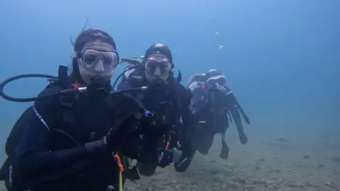 Rob Mason Three people are underwater wearing full diving and scuba-diving suits and equipment.