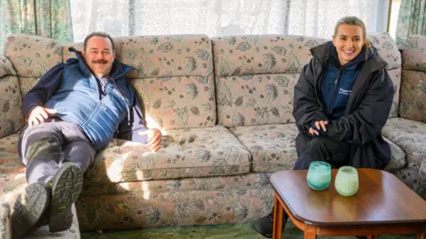 Sky UK/Ellis O’Brien The image shows two people sitting on a beige sofa with floral patterns sewed on it. There is a brown coffee table with two candles on it. Behind the couch is a window covered with white lace curtains.