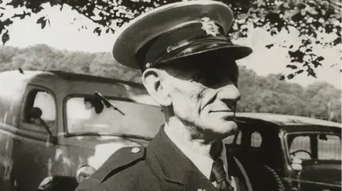Family handout George Randall, in a black and white image, wearing a uniform, with a hat on, jacket and tie and standing by two cars. He is also by a tree and is looking away to the right. 