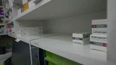 Un estante blanco con varias cajas de medicamentos. El resto del estante está vacío. Las cajas blancas y rectangulares más cercanas a la cámara llevan el nombre Dartacort en letras verdes y rosas.