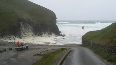 RNLI/Greg Spray An RNLI van looks onto Chapel Porth beach, surrounded by rugged cliffs, waves breaking in the background and an RNLI flag blowing in the wind.
