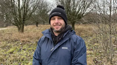 Martin Heath/BBC Sebastian Cummings with a short black beard smiling at the camera and wearing a black woolly hat and a blue jacket with a dark blue scarf. He is standing in an area of grass, scrub and trees.