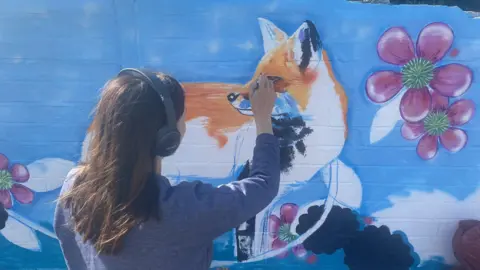 Chloe Farrant has her back to the camera - she has long brown hair and is wearing a long-sleeve blue top and headphones. With her right hand, she is painting the face of an orange and white fox on the wall, which has a blue background and purple and green flowers. 