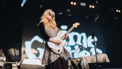 Nadine Ballantyne A blonde woman performing on stage. She is wearing dark sunglasses and a beige dress. She is playing a white electric guitar.