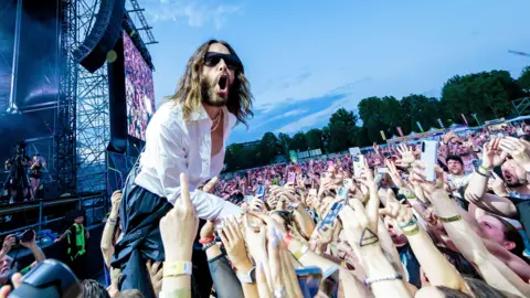 Getty Images Jared Leto performing at a concert. He wears a white shirt and is leaning into a sea of fans who have their arms raised up.