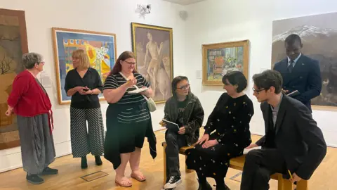 People in an art gallery are holding sketch books. Some are drawing. Others are sat listening to an artist tell them about the works they are studying.