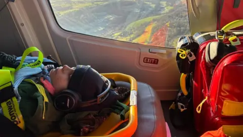 Inside a helicopter, the window in the background showing the landscape below. In the foreground, Dom, 12, lies unconscious on a stretcher with hat and coat and lots of straps to keep him in. There is a large bag of equipment to the right.