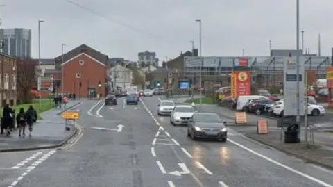 Google streetview of cars driving on King Street in Blackburn, with a garage on the right and flats on the left. People are walking on the pavement.