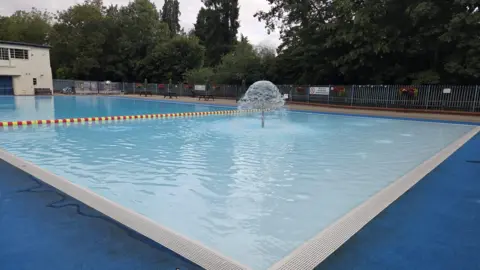 Seb Parker A poolside view of Droitwich Spa Lido. A dark blue edge runs around the pool while a single water fountain in positioned in the centre of the pool. A yellow and red rope runs across the width of the pool.
