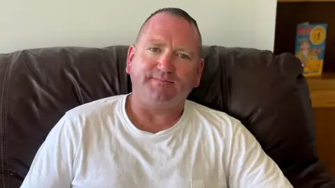 Michael Nicholson is seated on a brown leather armchair indoors, wearing a plain white T-shirt, facing the camera. Behind him is a light-colored wall with a set of electrical switches on the left and a wooden shelving unit on the right holding books and framed items. The scene is evenly lit with natural-looking light.