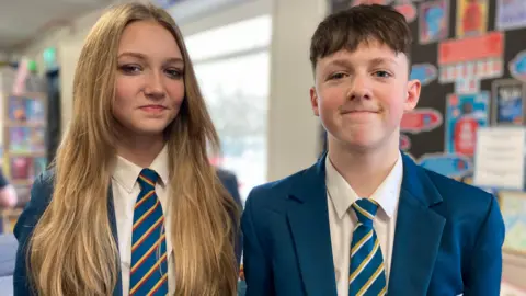 Ruby is standing on the left side of the image and harry is on the right. They are standing in a room with colourful posters on the walls. They are both wearing a blue blazer, white shirt and a blue tie with red and yellow stripes. 