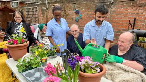 NIKKI FOX/BBC Workers at a care home help residents in an outdoor courtyard water plants on a table. The plants are in bloom and include hollyhocks and daffodils. They are yellow, purple and pink in colour. Four residents are visible, one is being helped to hold a green plastic watering can by one of the carers. He has a blanket over him.