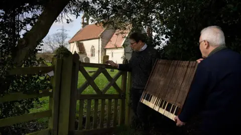 George Allan and assistant carrying part of an organ into his organ storage church. 