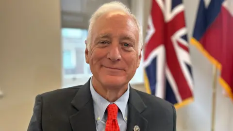 Kirk Watson, wearing a dark suit and red tie, stands indoors with a Union Jack flag and another flag visible in the background