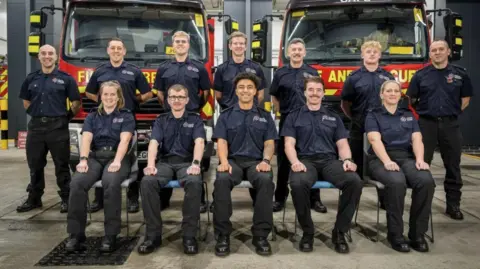 Hampshire and Isle of Wight Fire and Rescue Service A group of firefighters pose for a photograph in front of two parked fire engines. 