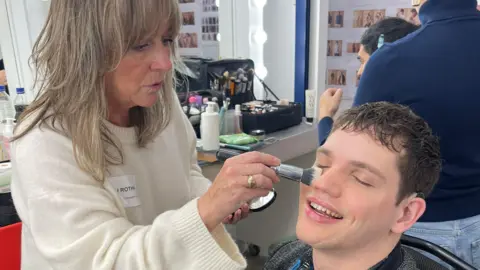 Chris Caswell A man, Elliot Caswell, is sitting down, having his makeup done in a dressing room by a makeup artist. He is wearing a grey jacket and a patterned shirt. He is smiling as the makeup artist applies powder to his face.