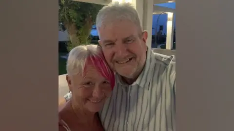 Family handout Tim McCarthy is wearing a white shirt with blue and green stripes has a grey beard and thick white hair. Hos wife has short blonde hair with a dyed plum coloured fringe.