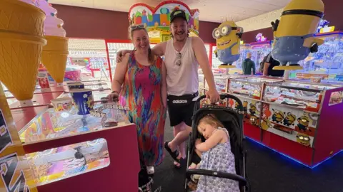 BBC / Kevin Shoesmith Stefan Careless, sporting a mullet hair style and wearing a vest top and shorts, is pictured with his partner Lucy Giacomin, who is wearing a multi-coloured long dress. Next to them is a pram containing their young daughter.
