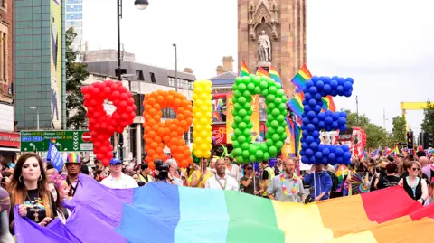 Pacemaker Press A large rainbow flag is being held by people taking part in a parade. The Albert clock is in the background. Pride balloons are being held up. 