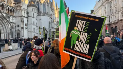 A shot of a crowd of Kneecap supporters outside the Royal Courts of Justice in London - a large Victorian Gothic Revival style in grey stone. The demonstrators are holding Irish tricolours and Palestinian flags. In the foreground is a placard showing a printed graphic design of a figure in a green and white tracksuit running. It reads "Free Mo Chara" in large white font.