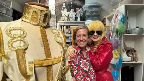 Two Rivers Media/BBC Scotland Kirsty Wark, in a red tile-patterned dress, is hugged by red PVC dress-wearing designer Pam Hogg, who is wearing striking yellow hair and sunglasses. A beige and sparkly military-style outfit is on a tailor's dummy in the foreground, a storeroom behind the pair, with shelves displaying platform shoes and many bags, boxes and drawers around.