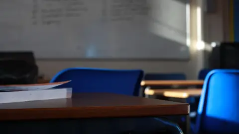 Getty Images A classroom. A brown desk with a book on it in the foreground, with blue chairs adjacent. Other desks and a white board can be seen in the background.