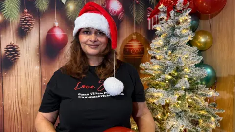 A woman has brown hair and is wearing a black top that says 'Susan Vickers Foundation'. She is wearing a Santa hat and there is a Christmas tree behind her.