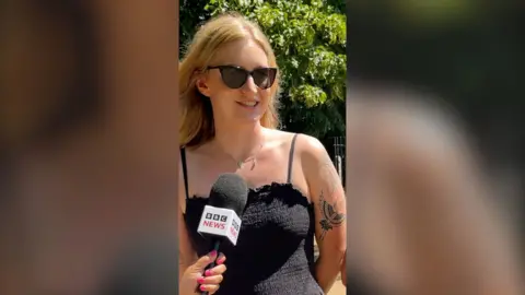 A woman speaking into a mic. She has blonde hair, sunglasses, a black strappy dress and tattoos.