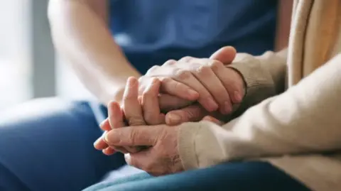 An elderly person's hands and a carer's hands placed on top of each other. The carer is wearing a blue uniform, the elderly person is wearing a beige-coloured top.
