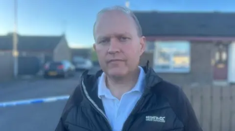 A man with balding hair, in a black jacket and white shirt, stood in front of bungalows and a police cordon tape