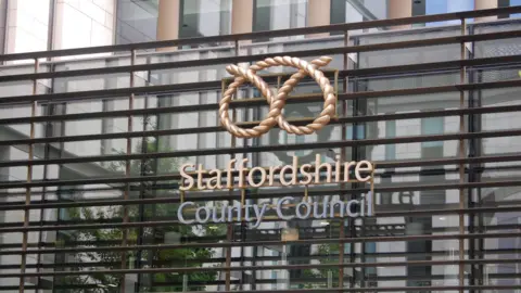 The front of a Staffordshire County Council building. It has the sign "Staffordshire County Council" on the front of the building which has glass with metal frames. 