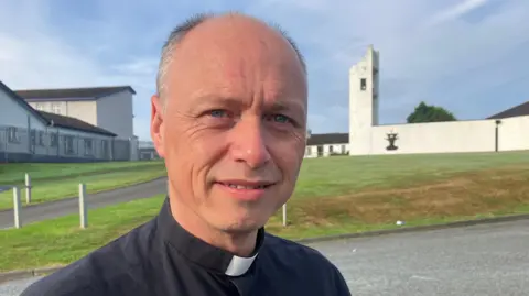 Fr Graham standing in front of a white church with a black shirt and white collar. He has a black shirt on and a white collar. He is partially bald with grey hair on the side and has blue eyes.