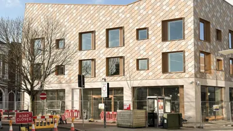 The new King's Lynn Library has distinctive cladding that looks a bit like dragon scales. It is coloured in shades of brown. In the foreground is a road closed sign.