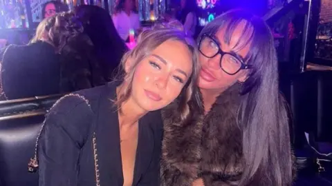Tallulah Bradford and her mother sitting together in a bar. Tallulah has her mid brown hair up behind her head, with curls on either side of her face, and is wearing a dark jacket. She is leaning on her mother, who has long dark brown hair, is wearing black-framed glasses and a brown fur jacket. 